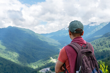 Naklejka premium male hiker from behind with backpack enjoys scenic mountain view, man is wearing cap , red shirt, standing against landscape of green hills and cloudy sky, adventure, connection with nature copy space