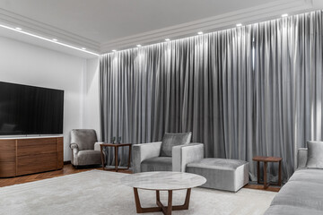 stylish large living room in white and gray tones, gray sofa, armchair, round table, wooden coffee table, carpet, floor lamp, gray curtains on windows. Illuminated by lamp. Space for relaxation