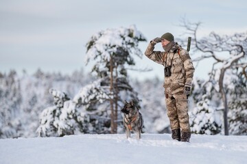 Hunter in winter camouflage with a dog in a snowy forest landscape, scanning the horizon