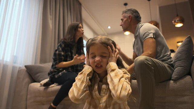 Desperate american 7-8 year old girl during parents quarrel closing ears at home. A little girl is sitting on the floor and covering ears with hands while mother and father arguing on background