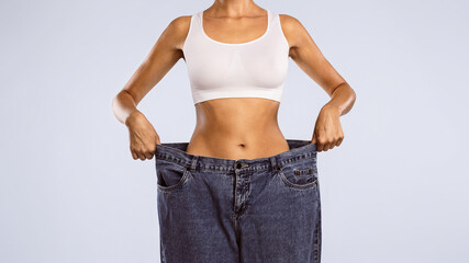 Closeup cropped view of skinny woman showing flat stomach pulling oversized big blue pants jeans. Slim body low fat. Healthy size athletic posing girl isolated over blue color studio background