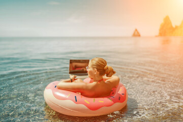 Woman laptop sea. Freelancer woman in sunglases floating on an inflatable big pink donut with a laptop in the sea. People summer vacation rest lifestyle concept