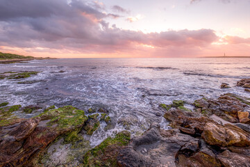 Serene Beauty Of A Rocky Shoreline At Sunset, With Sea Waves Gently Crashing Against The Rocks And A Lighthouse In The Background. The Sky Is Illuminated With Soft Pink And Orange Punta Prima, Menorca