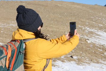 Woman hiker taking selfie photo using smartphone while hiking in winter jacket and clothing enjoying outdoor activity.