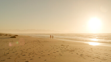 Two Silhouetted Figures Walk Along Beach By Waves At Sunset