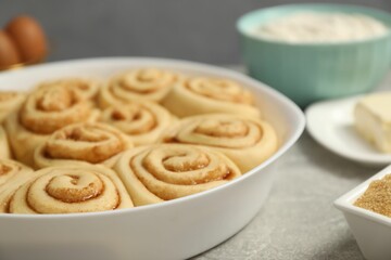 Raw cinnamon rolls in baking dish and ingredients on grey table, closeup