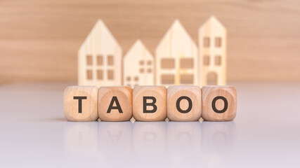 wooden blocks spelling TABOO with a blurred background of wooden houses on a reflective surface