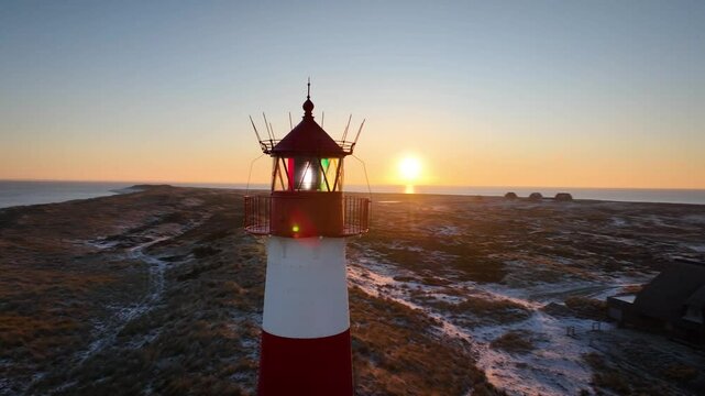 Aerial orbit of red and white striped lighthouse situated on a coastal landscape at sunrise in winter. Lighthouse West-List on the Ellenbogen (the "Elbow"), the peninsula on northern coast of Sylt isl