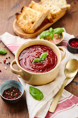 Tomato puree soup garnished with basil leaves and spices in ceramic bowl with toasted bread slices on wooden table