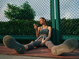 Young woman sitting on the ground checking her phone against a green mesh fence in a relaxed...