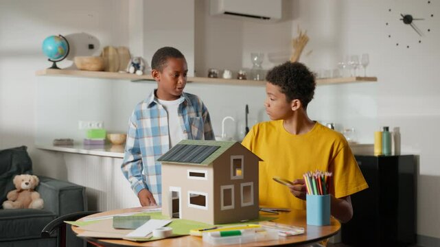 Older brother is already working on solar-powered model house, carefully assembling its parts. Younger brother enters frame, noticing progress and joining in to help, teamwork school science project