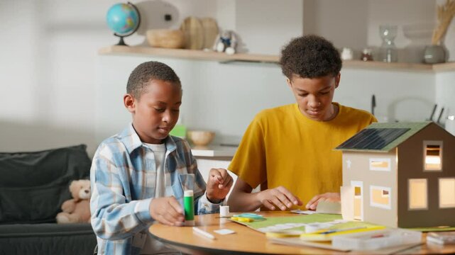 Two brothers celebrate success with a high-five after completing solar-powered cardboard house model for school science project. Scene highlights teamwork, creativity, and educational achievement.