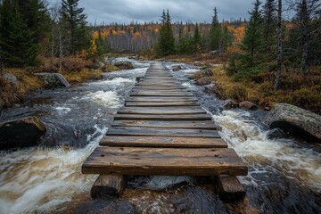 Obraz premium Rustic wooden bridge crossing a rushing river in a forest with autumn foliage, taken during overcast weather