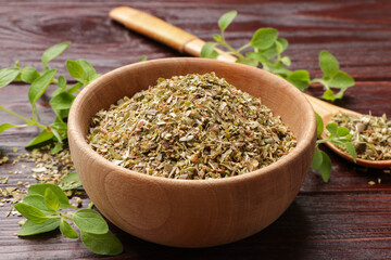 Dried oregano in bowl, spoon and green leaves on wooden table, closeup