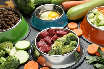 Natural and dry pet food in feeding bowls among products on dark textured table, closeup
