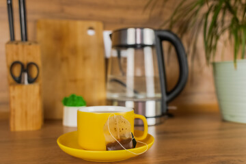 A yellow cup of tea stands on a yellow saucer and on the saucer lies a used tea poketik on a wooden table against the background of a teapot, a plant and kitchen utensils. The concept of tea drinking