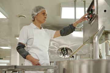 Woman operating industrial machinery in commercial kitchen, wearing hairnet and apron. She is ensuring proper functioning of the equipment, demonstrating focus and control