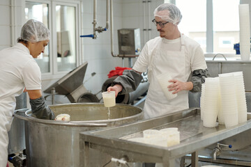 Workers wearing protective clothing in factory environment handling dairy products. Man and woman carefully pouring milk into containers next to processing equipment