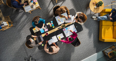Top View Shot: Diverse Entrepreneurs Sitting Around Conference Table in Tech Startup Office. Male and Female Colleagues Discussing Business Plan, Marketing, Using Laptop and Tablet Computer