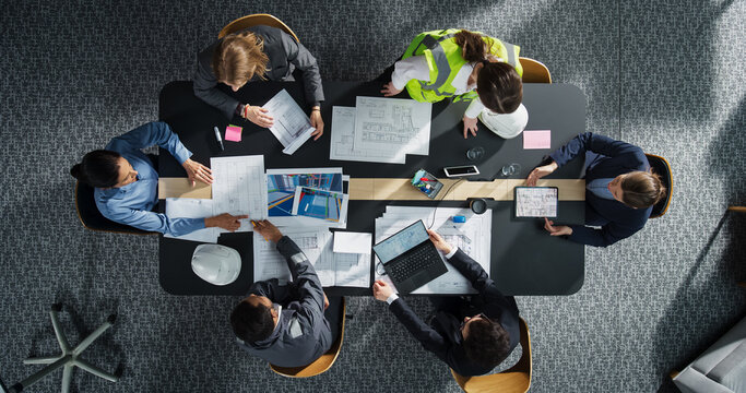 Top Down View of Diverse Team of Real Estate Construction Engineers Discussing Blueprints, Documents in Corporate Office. Modern Workspace of Architectural Bureau with Colleagues Using Tablet, Laptop