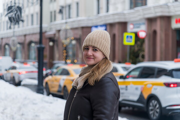 A young woman in a warm outfit stands on a snowy street, smiling at the camera. Yellow taxis are parked nearby, creating a lively urban atmosphere in winter.