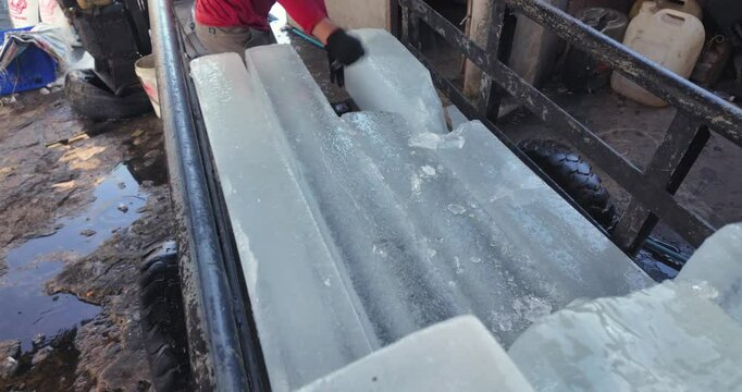 Man cuts a clear, rectangular block of ice with an icepick on a metal trailer, creating a spray of ice particles	