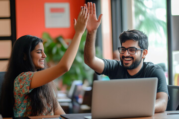 Smiling coworkers celebrating success with a high-five while working on a project in a vibrant, modern office with a laptop on the desk