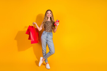 Young woman with blonde hair in trendy leopard print top holds shopping bags and phone against yellow background