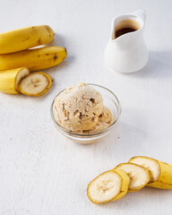 Banana ice cream is presented in a clear bowl alongside fresh banana slices and a small pitcher of syrup