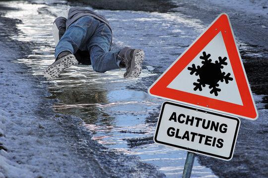 Schild mit Achtung Glatteis auf einem Gehweg im Winter. Ein Mann rutscht auf Glatteis aus