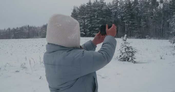 Passionate female photographer in winter clothes and glasses taking pictures of snowy forest landscape enjoying winter nature and photography. Woman taking pictures of fairytale winter forest.