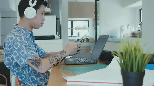 A focused young man wearing headphones is working on his laptop. He sits at a tidy desk, surrounded by study materials. The environment is bright and modern, highlighting a blend of productivity and l