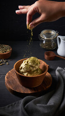 A hand sprinkles matcha powder over creamy matcha ice cream served in a wooden bowl. Various tea leaves and a milk jug add charm