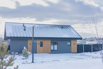 Modern house with a gray exterior and a wooden accent wall. The roof is covered in snow, a black fence surrounds the house. The house is located in a residential area. Contemporary country house