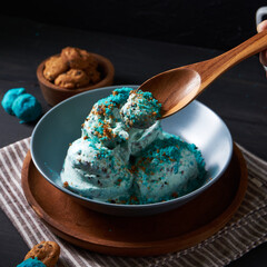 Creamy blue ice cream with cookie crumbles is served in a bowl, accompanied by a wooden spoon and chocolate chip cookies on the side
