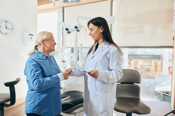Elderly woman shaking hands with female doctor holding clipboard, smiling, dressed in lab coat, both standing in bright medical office. Concept of healthcare and pensioners, medicine.