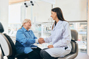Obraz premium Female doctor seated, shaking hands with smiling elderly woman in blue shirt seated in dental chair, dental equipment in bright office against white background. Concept of healthcare and pensioners.