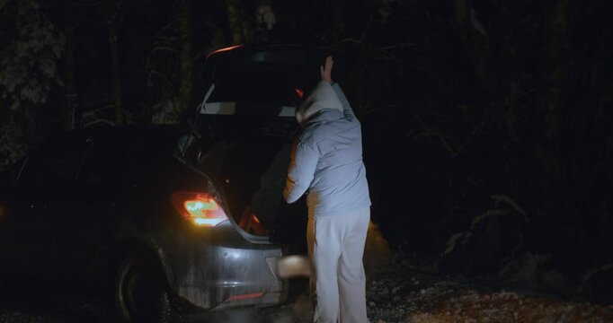On a dark winter night, a woman retrieves a warning triangle from her car's trunk and positions it on a snowy road behind her vehicle, indicating a breakdown in a remote forest area.