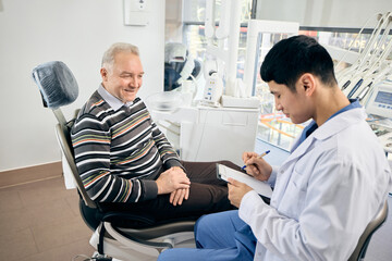 Dentist, holding clipboard, discusses dental implants with patient reclined in chair, sunlight streaming into modern, well-equipped office. Concept of healthcare and pensioners, medicine.