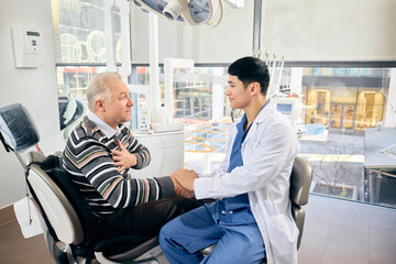Fototapeta premium Dentist holds hand of patient in chair, offering comfort in bright, modern dental office, large windows behind. Concept of healthcare and pensioners, medicine, treatment.