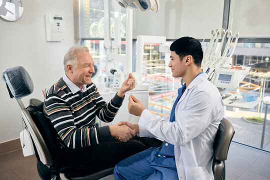 Smiling dentist shakes hand of laughing old man, patient in chair, both gesturing, friendly atmosphere in bright, modern dental office. Concept of healthcare and pensioners, medicine.