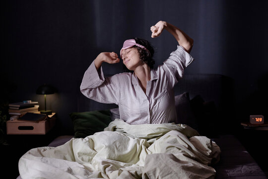 Wide shot of brunette curly haired woman doing morning stretching after sleep while sitting in bed and enjoying warm of sunlight