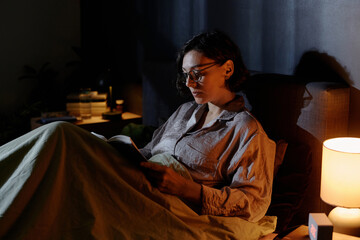 Brunette woman with bob hair cut resting in bed with hovel in hands, she wearing round glasses to...