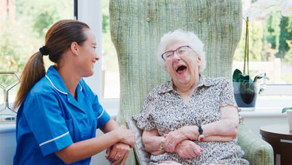 Laughing Senior Woman Sitting In Chair In Residents Lounge And Talking With Nurse In Retirement Home