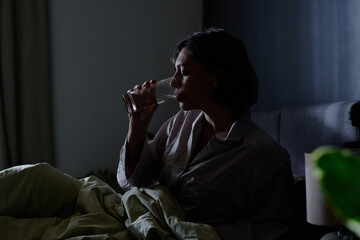 Woman in pajamas sitting in bed in middle of night and taking sip of pure water