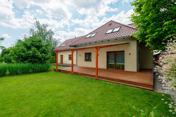 Modern house with a red tiled roof, spacious wooden deck with a pergola, and a lush green backyard surrounded by trees and landscaping on a sunny day.