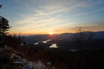 Golden hour sunlight bathes a tranquil mountain valley and lake, creating a picturesque scene of nature's beauty during twilight with colorful sky and silhouettes of trees and hills