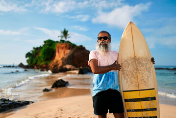 Senior man with beard holds surfboard on sunny beach. Elderly surfer smiles, enjoys active retirement on ocean shore. Happy mature male embraces vibrant lifestyle. Summer adventure, freedom.