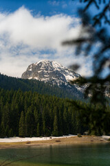Snow capped mountain peak rises above a dense coniferous forest bordering the tranquil black lake,...