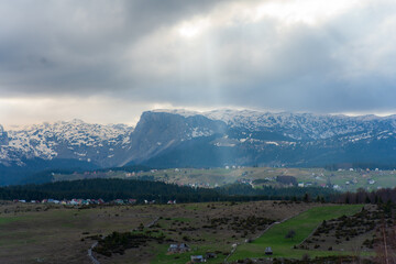 Fototapeta premium Sun rays are illuminating snowy mountain peaks over a green valley with scattered houses and coniferous trees, creating a picturesque landscape in montenegro